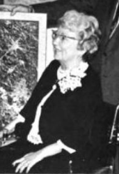 An older white woman wearing a dark suit and glasses, seated in a wheelchair. She is smiling, and wearing a corsage. One hand is resting on her lap.