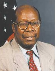 A portrait photo of a balding black man in front of a US flag; he is wearing spectacles, a tan suit, red tie, and is looking to the camera's right.