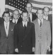 Six standing men in suits and ties, with 48-star American flag covering the wall behind them.