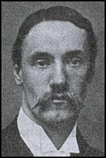 A head and shoulders view of a dark-haired man with a moustache, wearing a suit jacket and wing collared shirt