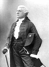 Three-quarters monochrome photograph of a man facing to his right, dressed in a white wig, ruffled shirt, high-waisted trousers with sword hanging from the belt, jacket, and gloves. He rests his right hand on a walking stick, and holds a large Napoleon-style hat under his left arm.