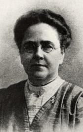 A middle-aged white woman wearing glasses and a high-necked blouse; her hair is arranged in a slightly bouffant top bun.