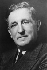 A middle-aged man wearing a coat and tie looks into the camera.