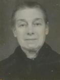 a black and white photograph of an older white woman with her hair in a bun, wearing a black coat and facing the camera head on
