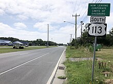 A four-lane asphalt road with a grassy median surrounded by development.