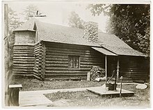 A large log cabin-like building, with a chimney, window with curtains, a small porch, and a water pump out front. The building is rectangular with a round room in the back