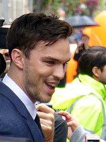 A young, Caucasian man with short, dark hair and facial stubble wearing a blue jacket and white shirt against a busy, outdoor background. Behind him is a man in a high-visibility jacket and a woman with an umbrella.