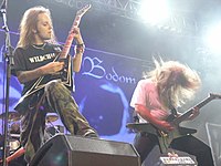 A color photograph of two members of the group Children of Bodom standing on a stage with guitars, drums are visible in the background