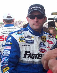 Scott Wimmer in his blue racing suit with a white chest filled with sponsors, wearing sunglasses and a black Fastenal baseball cap