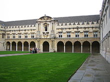 Facade of an ancient building, facing an expanse of grass. At ground floor level the building presents a long colonnade of arches, above which are rows of windows up to a crenellated roof line.