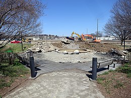 Construction equipment has begun to dig up concrete and level the earth beyond an area with a concrete fountain and wooden benches.