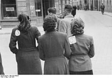 Schwarzweißfotografie von drei Frauen, die der Kamera ihren Rücken zugedreht haben. Die linke und rechte Frau haben an ihrer linken Schulter einen Zettel mit einem eingekreisten J geheftet. Sie laufen in einer Straße hinter zwei Männern.