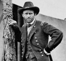 Photograph of Grant, staring at the camera while standing next to a tree