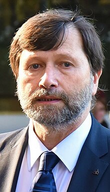 white male with salt-and-pepper beard, wearing suit and tie, looking at camera