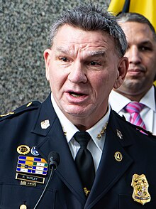 Worley, an older man with black-gray hair, wearing a black suit jacket with a nametag, the badge of the Baltimore Police Department, and various other badges, over his white police uniform and a black tie, giving a speech.