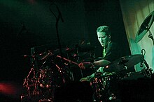 Drummer with crewcut, wearing black T-shirt, intently plays his large drum set in the spotlight.