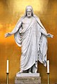 Frontale Farbfotografie von einer Jesusfigur in weißem Marmor, die auf einem Podest steht und die Arme ausbreitet. Sie trägt eine Toga und steht vor einer goldenen Wand. Vor ihr stehen zwei Kerzenhalter.