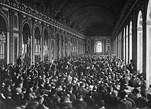Photograph of the long Hall of Mirrors where an immense crowd is packed standing up around a group of seated individuals.
