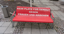 Das Foto zeigt eine Sitzbank, die rot bemalt ist und auf der Lehne in Versalien "KEIN PLATZ FÜR GEWALT GEGEN FRAUEN UND MÄDCHEN" anzeigt