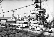 Two large warships lie in port; several men can be seen on the deck of the closer vessel, while the further one has a small boat tied alongside.