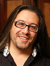 Face of a smiling man with long black hair and glasses