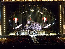 AC/DC shown on stage with huge screens either side depicting Angus on guitar. He is actually at extreme stage right the rest of the band at centre stage. They are beneath a huge steam engine, tilted slightly upwards, with AC/DC's logo on its side.
