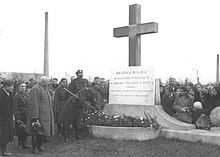 A large group of people, many of them in Polish Army winter uniforms, surround a monument. The monument consists of a concrete base and a 5-metre-high cross, with a large stone tablet in front of it. The text on the tablet reads (in Polish): Brotherly grave of heroes fallen in defence of the fatherland in 1920; the monument was funded by workers of the Polish Bank in Warsaw. In the background a tall factory chimney.