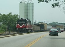 A freight train is seen on a track parallel to a road with cars on it. The train is in two sections. While on section with one locomotive is sitting on the main line, the other section is in a siding lead by the other locomotive, switching a local industrial customer served by the railroad.