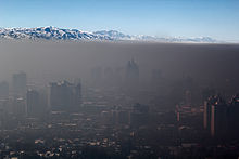 Dense, smokey air lays over a cityscape like a blanket. In the distance, a mountain range and clear blue sky can be seen.