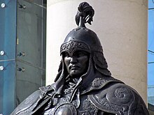 Torso and head of a statue of a man in full ceremonial armour with plume on helmet, in front of a pillar.
