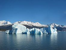Fotografie eines Eisberges. Im Vordergrund eine Wasseroberfläche, im Zentrum ein hellblauer Eisberg. Im Hintergrund schneebedeckte Berggipfel und ein wolkenloser Himmel.