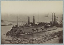 A ship along a shoreline. The vessel has two smokestacks, flies the American flag, and multiple cannon are visible.