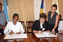 Photograph of two seated women, attended by two witnesses standing beside them, signing documents on a table.