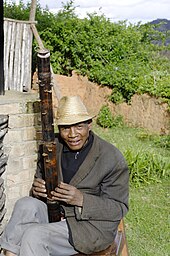 An elderly man sitting with a valiha instrument on his knees in front outside a house