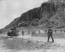 tank in mid-left heading down road from left to right. Terrain is flat in foreground, forested cliffs in back ground. Soldiers are walking behind the tank