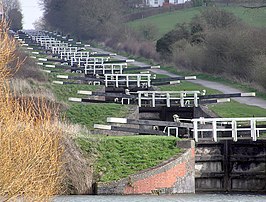 A series of approximately 20 black lock gates with white ends to the paddle arms and wooden railings, each slightly higher than the one below. On the right is a path and on both side's grass and vegetation.