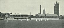 Black and white photograph of a cricket ground with players on the field dressed in white. In the background two churches, a tall industrial chimney and some houses can be seen.