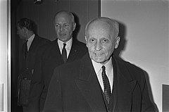 Panagiotis Pipinelis and another man, both in dark suits, walk through a doorway toward the camera