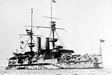 A large gray ship with blocky sides, two tall masts, and two funnels sits at anchor