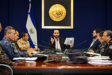 Nayib Bukele seated at the head of a table holding a piece of paper in his left hand in front of various cabinet officials