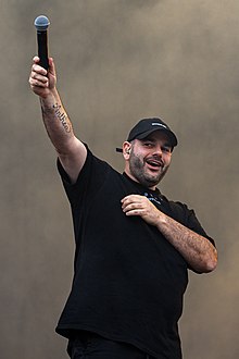 A full-bodied man with dark stubble and armhair in all-black casual garb (including baseball cap) holds a mic up high and smiles slightly while he performs.