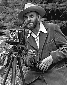 A photo of a bearded Ansel Adams with a camera on a tripod and a light meter in his hand. Adams is wearing a dark jacket and a white shirt, and the open shirt collar is spread over the lapel of his jacket. He is holding a cable release for the camera, and there is a rocky hillside behind him. The photo was taken by J. Malcolm Greany and first appeared in the 1950 Yosemite Field School Yearbook.