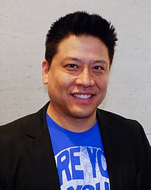 An image of a man with short, black hair and a blue shirt and black jacket. He is smiling toward the camera.