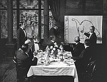 A black and white film still. A group of men sit around a dining table in the center. To the right, a man stands by and gestures at a large drawing of a dinosaur.