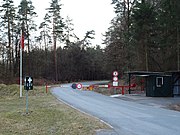 Guardpost with vehicle access control barrier, and red 'danger' flag aloft its flagstaff. The circular signs on the pole to the right indicate 'no pedestrians' (top) and 'no vehicles' (bottom).