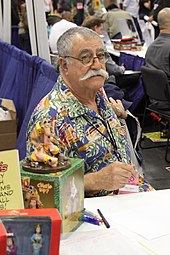 A man with thinning, greying hair and a thick, white moustache sits behind a table. He wears a colourful shirt. Pens and action figures of his works are on the table in front of him.