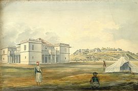A white mansion and, in the distance, a fortified palace atop a hill. Three men stand near a tent in the foreground. The mansion has a large rust-coloured shingled roof, pillared porches and open-air balconies on its four sides, and large box-like corner sections that extend out from the rest of the architecture.
