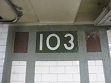 Tiles on the walls of the platform extension. There are vertical and horizontal green mosaic bands, surrounding white ceramic bricks on the bottom and three rectangular mosaic boxes on the top. The center box contains the number "103" in white tiles on a green background. The left and right boxes contain bronze tiles.