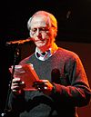 An elderly and spectacled Don DeLillo is pictured reading from paper at a microphone. He is wearing a sweater over a collared shirt.