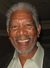 An African American man with a beard and short hair, both a mix of white and grey, and wearing an earring in each ear: He is smiling towards the camera.